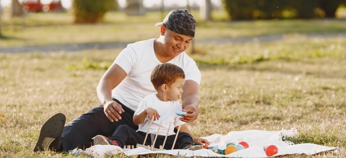 A joyful father and son playing together on a blanket in a sunlit park.